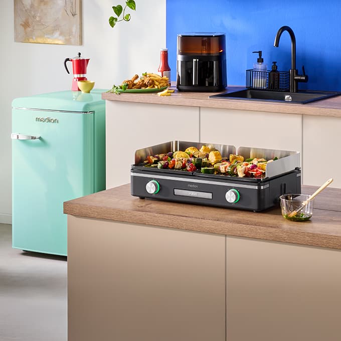 Tischgrill von MEDION auf Kücheninsel mit gegrilltem Gemüse und Spießen. Im Hintergrund weitere Küchengeräte in moderner Küche.