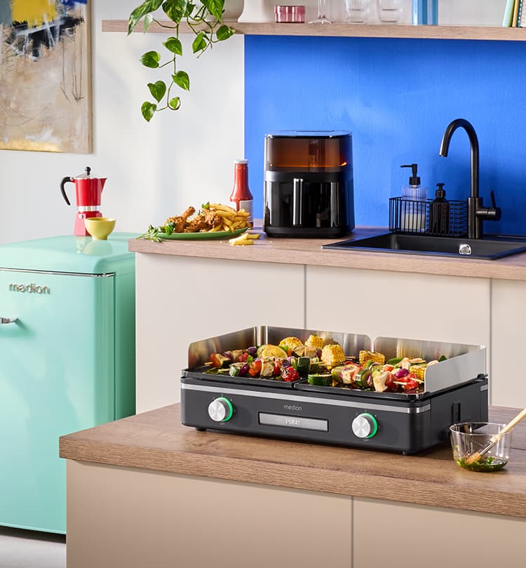MEDION Tischgrill mit gegrilltem Gemüse auf einer Küchenarbeitsplatte. Im Hintergrund Heißluftfritteuse, Spüle und Küchendekoration in moderner Küche.