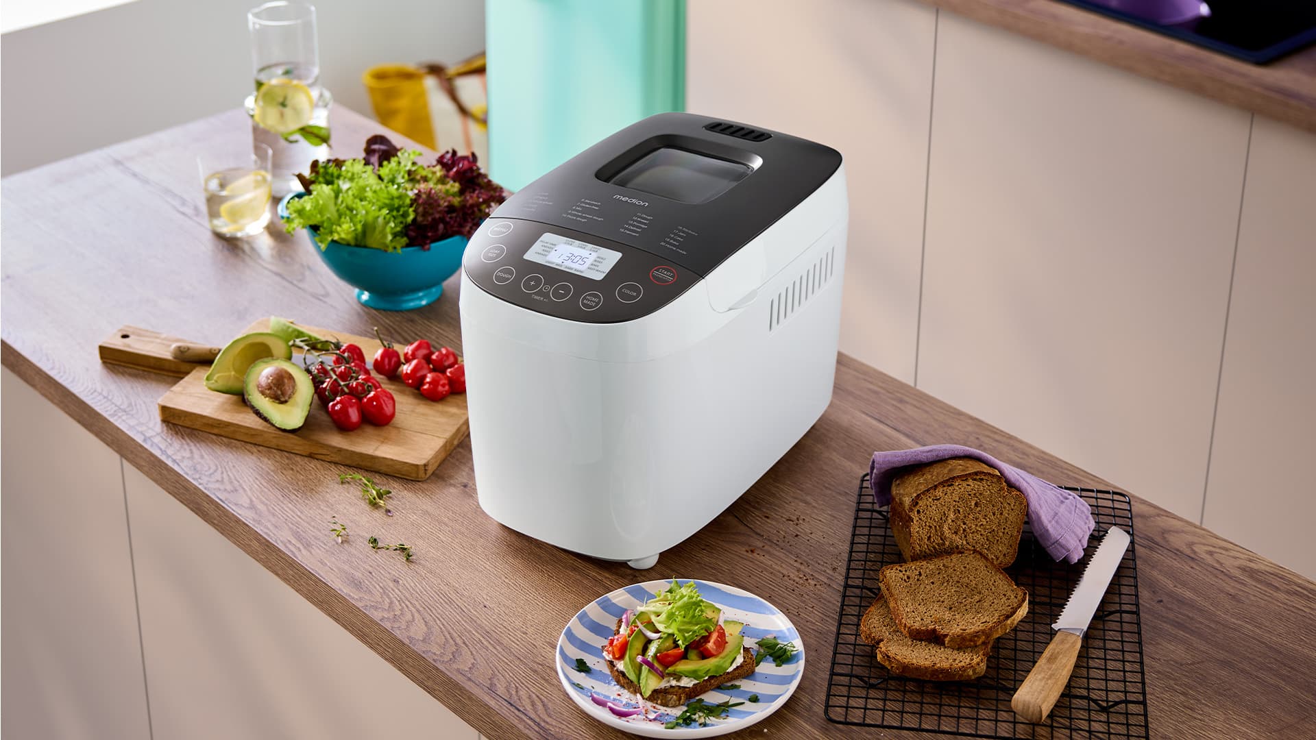 Brotbackmaschine auf einer Küchenzeile. Ein angeschnittenes Brot, ein Brett mit Avocado und Cherrytomaten und eine Schlüssel mit Salat stehen daneben