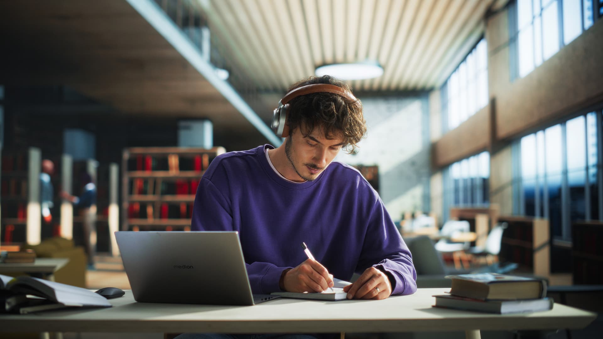 Ein Student in der Bibliothek am Schreibtisch sitzend mit einem Notizblock und einem Stift in der Hand, schreibend. Vor im steht ein aufgeklapptes Laptop.