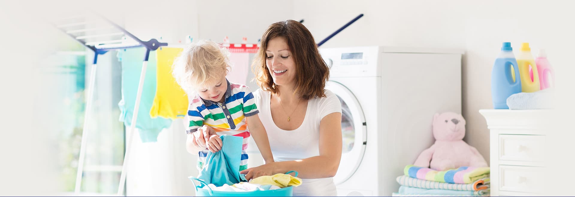 Frau und Kind waschen Wäsche in einem hellen Raum; im Hintergrund sind eine Waschmaschine, ein Wäscheständer und bunte, gefaltete Handtücher zu sehen.