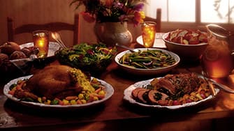 Ein Tisch mit gebratenem Hähnchen, Aufschnitt, Salaten, grünen Bohnen, Brötchen und Eistee in einem warm beleuchteten Speisesaal.