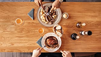 Zwei Personen speisen an einem Holztisch mit Tellern voller Fleisch, Kartoffeln, Brot und Getränken. Luftaufnahme, die Hände mit Besteck zeigt.
