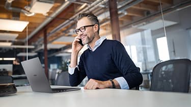 Ein Geschäftsmann mit der rechten Hand telefonierend mit einem Handy. sitzend an einem Schreibtisch. Vor ihm steht ein aufgeklapptes Handy. Die ganze Szenerie wird schräg von der rechten Seite gezeigt.