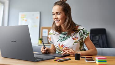 Eine lachende Frau sitzt am Schreibtisch und zeichnet mit ihrem Grafiktablett. Ein aufgeklapptes Laptop liegt vor ihr.