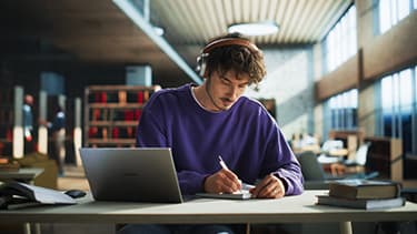 Ein Student in der Bibliothek am Schreibtisch sitzend mit einem Notizblock und einem Stift in der Hand, schreibend. Vor im steht ein aufgeklapptes Laptop.