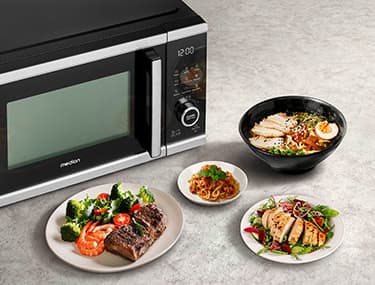 Mikrowelle neben Tellern mit gegrilltem Steak mit Gemüse, Ramen, Pasta und einem Salat mit Hähnchenscheiben auf einer hellen Arbeitsplatte.