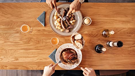 Zwei Personen speisen an einem Holztisch mit Tellern voller Fleisch, Kartoffeln, Brot und Getränken. Luftaufnahme, die Hände mit Besteck zeigt.