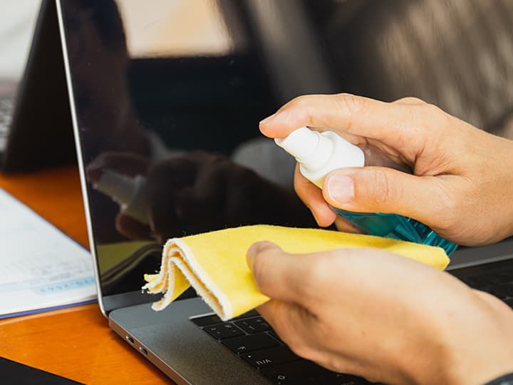 Hände reinigen einen Laptop-Bildschirm mit einem gelben Tuch und einer Sprühflasche auf einem Holzschreibtisch, auf dem teilweise ein Papier sichtbar ist.