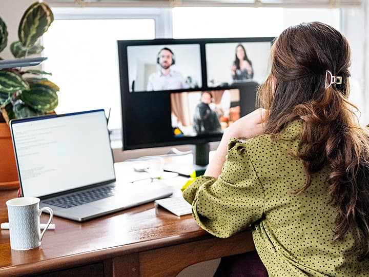 Frau in grüner Bluse bei einem Videoanruf an einem Schreibtisch mit einem geöffneten Laptop und einem Monitor mit vier Personen