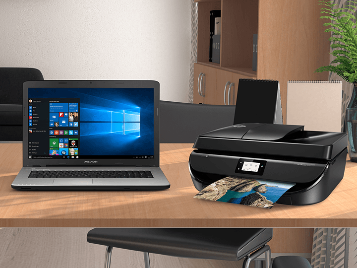 Ein Laptop mit Windows 10 und ein Drucker mit einem ausgedruckten Foto stehen auf einem Holzschreibtisch in einer modernen Büroumgebung.