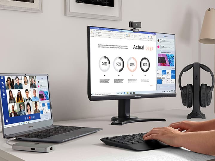 Eine Person arbeitet an einem Schreibtisch mit einem Laptop und einem Monitor, auf dem Diagramme und ein Videoanruf angezeigt werden. Kopfhörer hängen in der Nähe an einem Ständer.