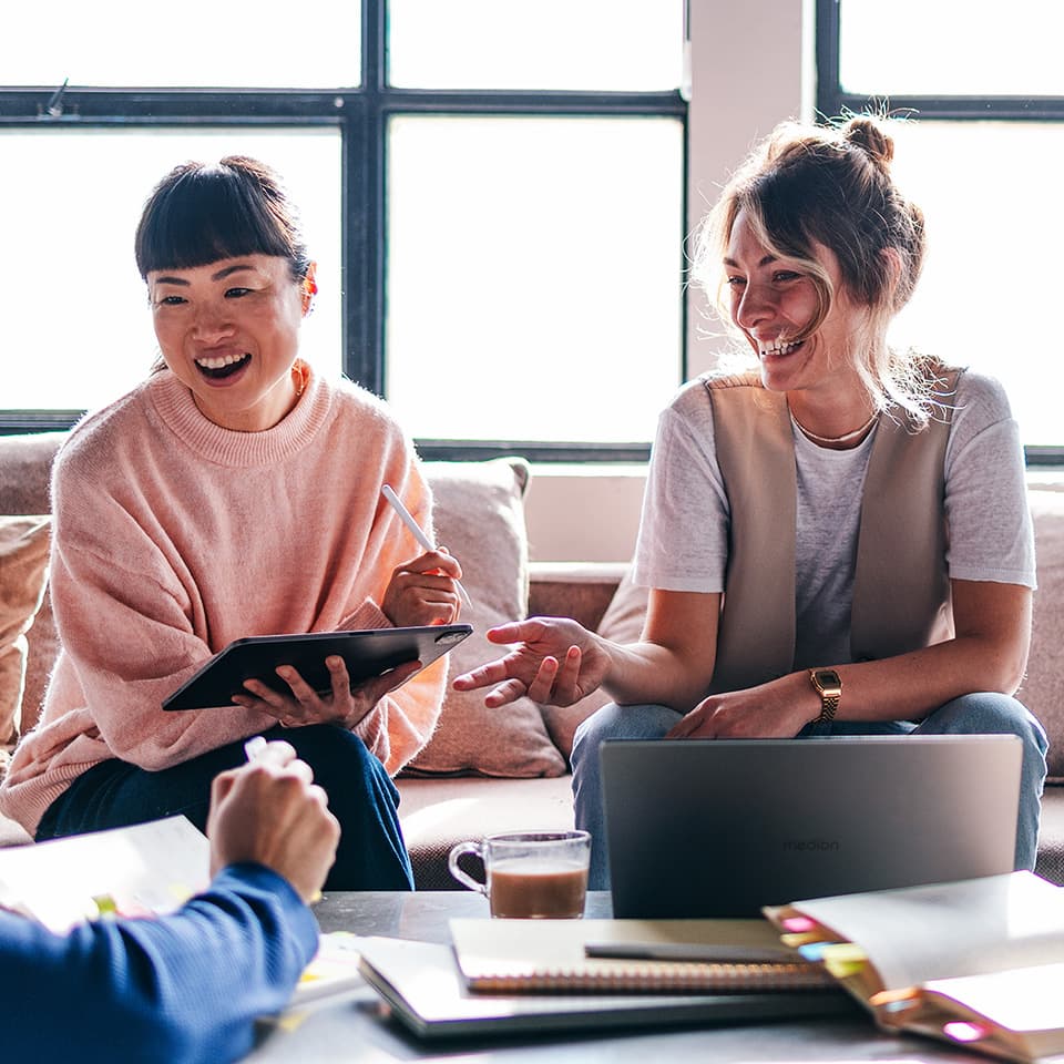 Zwei lachende Frauen, die nebeneinander sitzen und sich unterhalten. Ein Laptop und ein Tablet kommen dabei zum Einsatz.