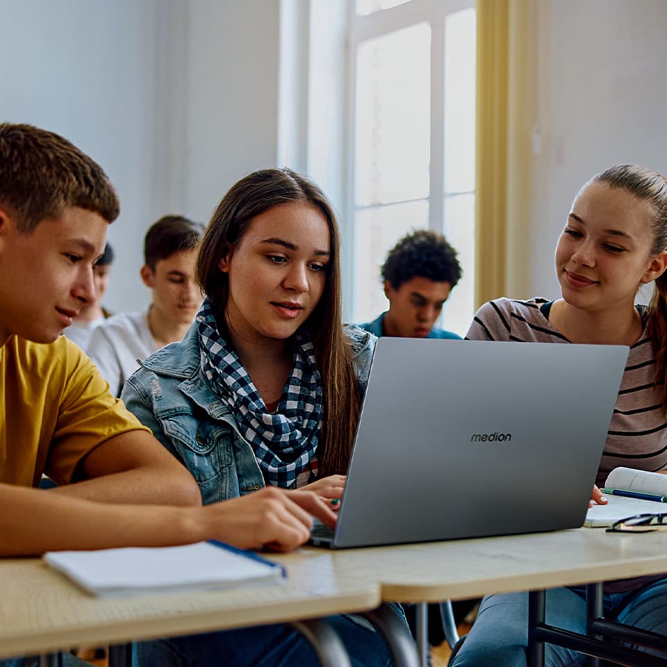 Eine Gruppe von Schülern im Klassenraum. Das Mädchen sitzt vor dem aufgeklappten Laptop. Links und rechts von ihr sitzen zwei Mitschüler.