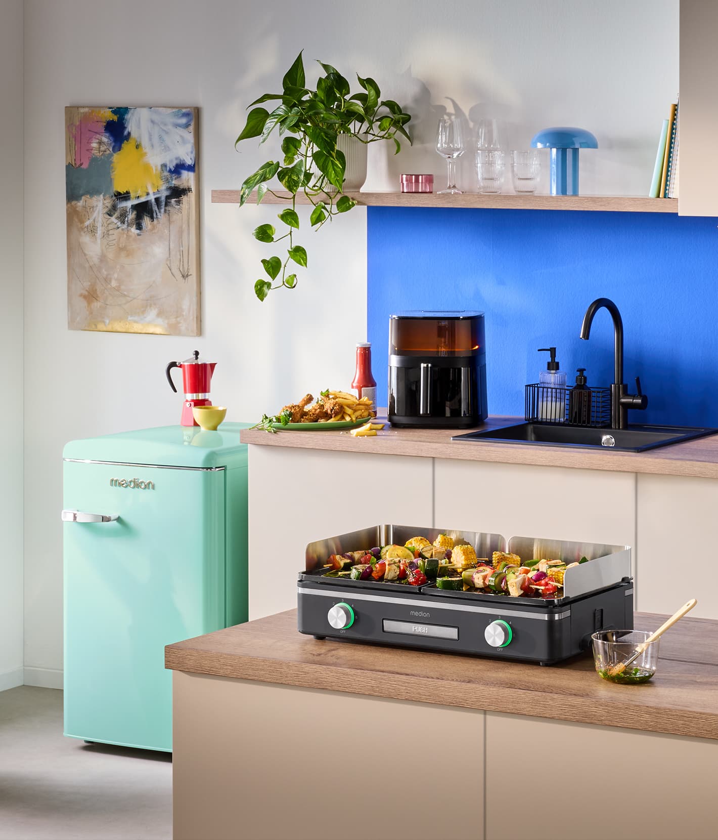 Moderne Küche mit verschiedenen MEDION Küchengeräten, darunter Tischgrill mit Gemüse und Spießen, Heißluftfritteuse, Küchenmaschine, Mikrowelle und mintfarbener Kühlschrank. Helle, wohnliche Küchenszene.