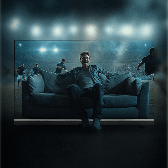 Großer Fernseher zeigt einen Zuschauer auf einem Sofa in einem Fußballstadion, während Spieler im Hintergrund auf dem Feld laufen. Eindrucksvolle Stadionatmosphäre mit Flutlicht.