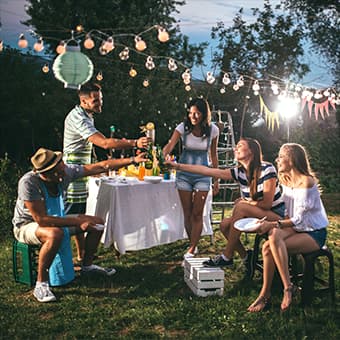 Eine Gruppe von Freunden genießt eine Gartenparty im Freien. Sie sitzen und stehen rund um einen Tisch mit Getränken, umgeben von Lichterketten und Dekorationen.