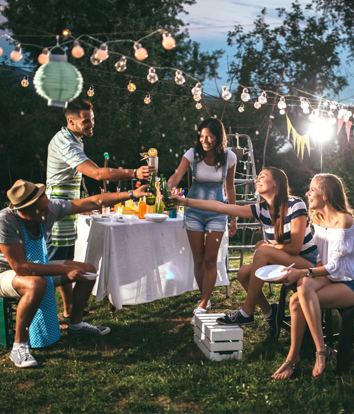 Eine Gruppe von Freunden genießt eine Gartenparty im Freien. Sie sitzen und stehen rund um einen Tisch mit Getränken, umgeben von Lichterketten und Dekorationen.