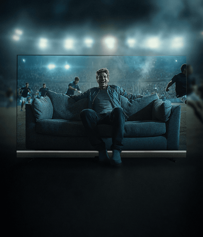 Großer Fernseher zeigt einen Zuschauer auf einem Sofa in einem Fußballstadion, während Spieler im Hintergrund auf dem Feld laufen. Eindrucksvolle Stadionatmosphäre mit Flutlicht.