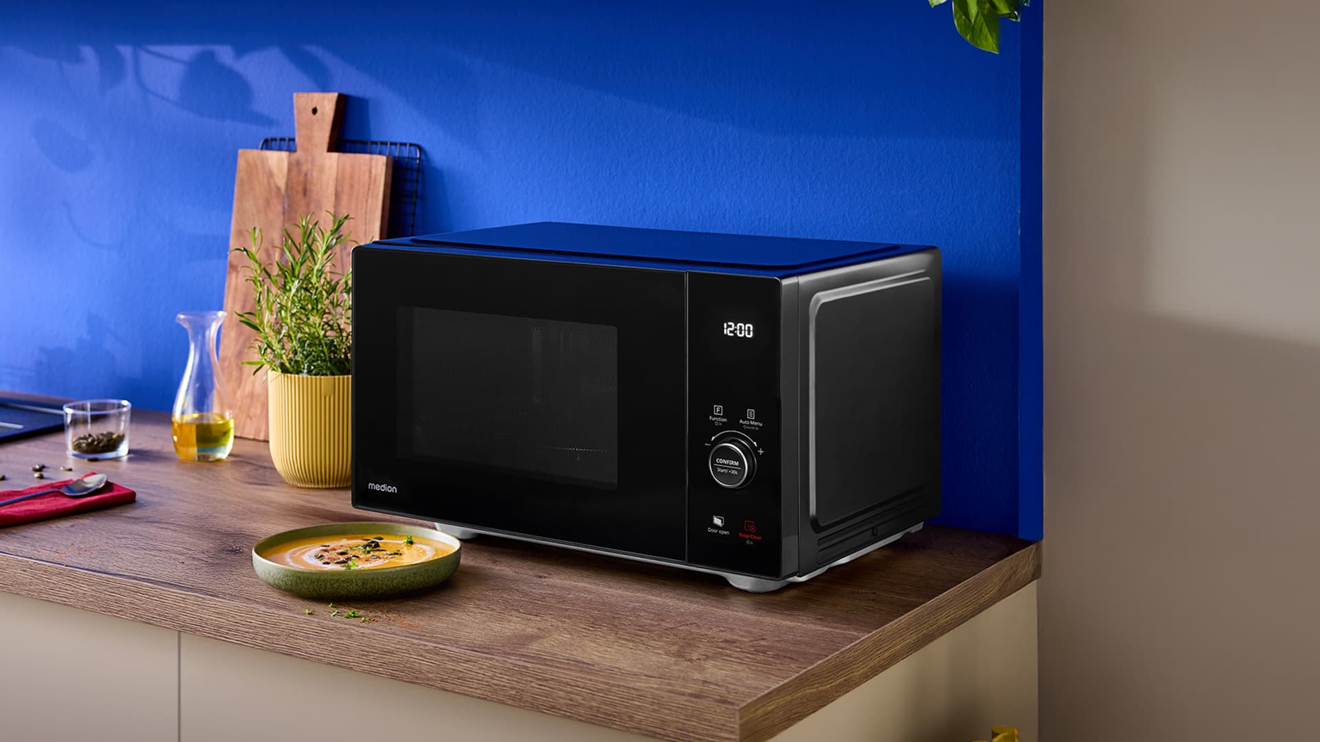 Eine schwarze MEDION-Mikrowelle auf einer hölzernen Küchenarbeitsplatte vor einer blauen Wand, umgeben von einem Schneidebrett, Kräutern und einem Teller mit Essen.