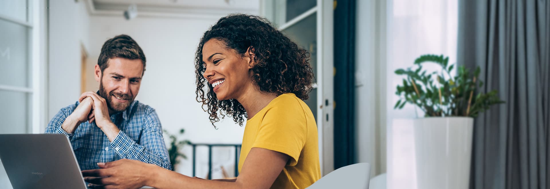 Zwei Personen sitzen gemeinsam vor einem MEDION Laptop in einem hellen Raum und lächeln – im Hintergrund steht eine große Zimmerpflanze.