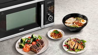 Mikrowelle neben Tellern mit gegrilltem Steak mit Gemüse, Ramen, Pasta und einem Salat mit Hähnchenscheiben auf einer hellen Arbeitsplatte.