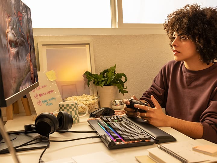 Person mit lockigem Haar spielt an einem Schreibtisch ein Videospiel und hält einen Controller in der Hand. In der Nähe befinden sich Kopfhörer, eine Tastatur und eine kleine Pflanze.