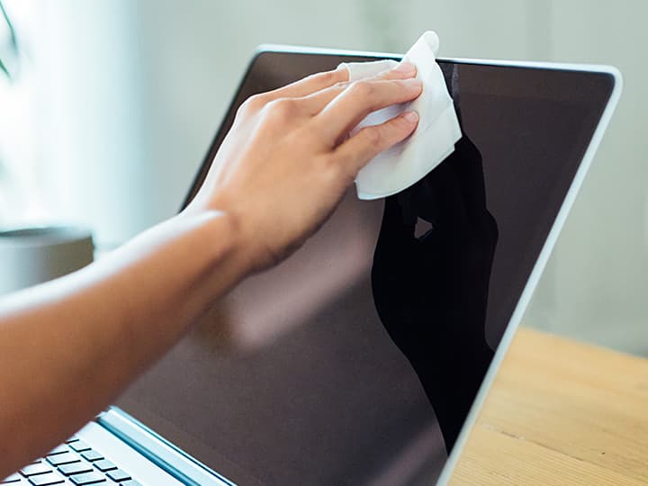 Reinigen eines Laptop-Bildschirms mit einem weißen Tuch von Hand auf einem Holzschreibtisch.