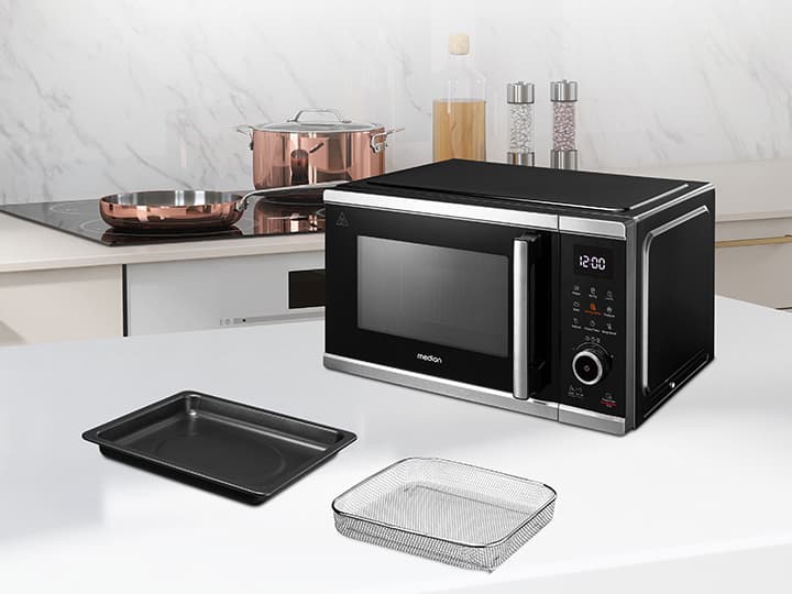 Eine schwarze Mikrowelle auf einer weißen Arbeitsplatte mit Backblech und Glasschale. Im Hintergrund stehen Kupfertöpfe und Gewürze.