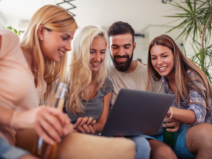 Vier lächelnde Menschen schauen gemeinsam in einem gemütlichen Raum mit Pflanzen auf einen Laptop-Bildschirm. Das geöffnete Laptop liegt auf dem Schoß einer Frau.