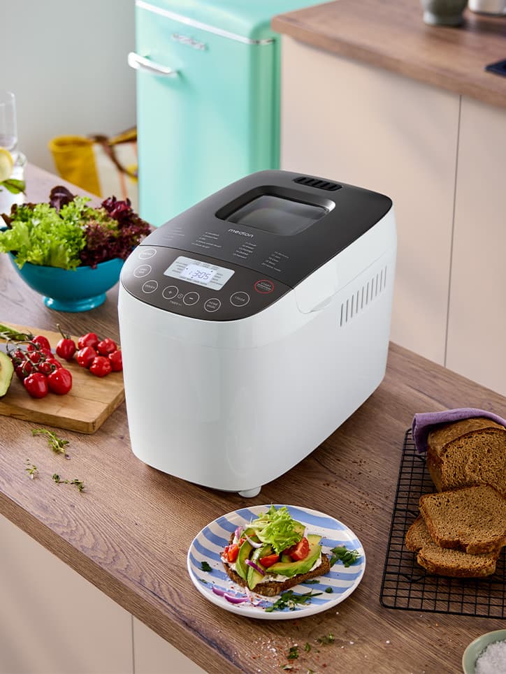 Brotbackmaschine auf einer Küchenzeile. Ein angeschnittenes Brot, ein Brett mit Avocado und Cherrytomaten und eine Schlüssel mit Salat stehen daneben