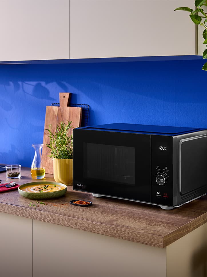 Eine schwarze MEDION-Mikrowelle auf einer hölzernen Küchenarbeitsplatte vor einer blauen Wand, umgeben von einem Schneidebrett, Kräutern und einem Teller mit Essen.