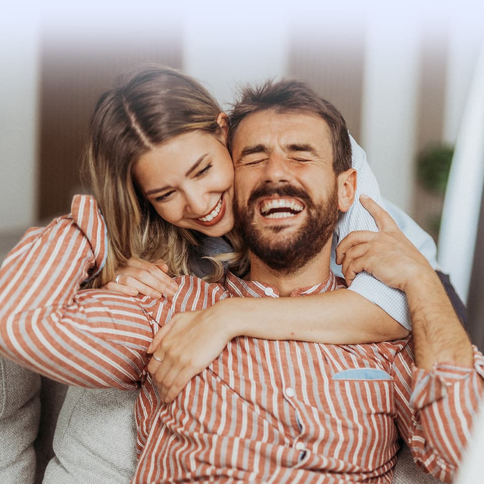 Eine Frau umarmt einen lachenden Mann von hinten auf einem Sofa. Beide tragen gestreifte Hemden und wirken in der gemütlichen Umgebung fröhlich.