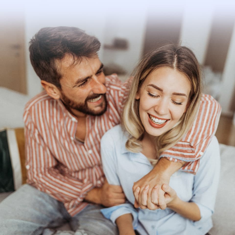 Ein lächelndes Paar sitzt zusammen, der Mann im gestreiften Hemd umarmt die Frau von hinten im hellblauen Hemd, beide lachen fröhlich.