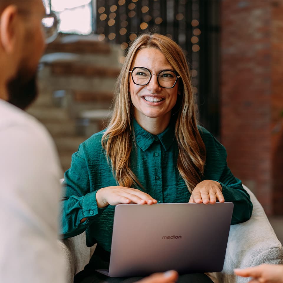 Eine lachende Frau, die auf ihrem Schoß ein aufgeklapptes Laptop positioniert hat. Sie schaut zu einem Mann, der mit dem Rücken zum Betrachter zu sehen ist.